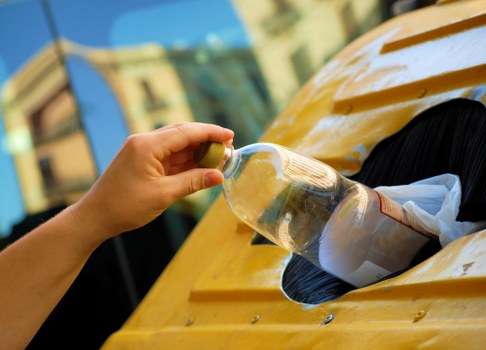 A person’s hand is seen placing a clear plastic bottle with a label into a circular opening on the side of a yellow waste collection bin, which is positioned outdoors on a paved surface. The background shows blurred buildings and a bright sky, indicating a residential or urban setting. The bin’s surface appears to be smooth with a glossy finish, and the opening is designed to accept bottles or similar recyclable materials. The scene suggests an active process of rubbish disposal, emphasizing waste separation or collection typical of a domestic or commercial rubbish removal service in Richmond. The image illustrates proper recycling practices as part of garden or property clearance, relevant to services such as those provided by Garden Clearance Richmond. The environment is well-lit with natural daylight, and the focus is on the transfer of the bottle, highlighting the action of rubbish collection or disposal activity.