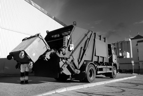 A large commercial rubbish collection truck is positioned on a paved street outside an industrial or warehouse building, with its rear loading mechanism engaged. The truck's metal body features a smooth, reflective surface, and the rear section is open, revealing a partially lifted hydraulic lift arm attached to a large, wheeled waste container. The container, made of sturdy metal with a textured finish, is tilted forward, indicating it is in the process of being emptied or loaded. A worker wearing work trousers and safety gloves stands nearby, assisting with the rubbish collection process. The background includes a tall, corrugated metal wall on the left and additional industrial buildings with ventilation units and silos visible in the distance, against a partly cloudy sky. The scene is captured in natural daylight, highlighting the industrial setting typical for commercial rubbish removal services in Richmond and surrounding areas.