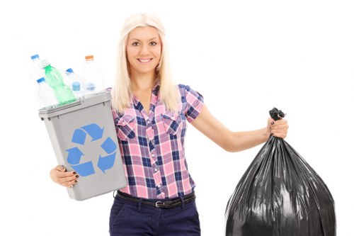 A smiling woman with long blonde hair, wearing a short-sleeved checkered shirt in shades of blue and pink, is standing against a plain white background. She holds a small grey recycling bin filled with clear plastic bottles and a couple of plastic containers in her left hand, with the recycling symbol visible on the front of the bin. In her right hand, she is gripping a tightly tied large black plastic rubbish bag. The scene emphasizes waste removal and recycling, consistent with services such as those offered by Garden Clearance Richmond. This presentation of waste collection includes details such as the transparent plastic bottles inside the recycling bin, the textured surface of the plastic bag, and the casual attire of the woman, all suggesting a positive and organized approach to rubbish management, suitable for a residential or small commercial setting in or near Richmond.