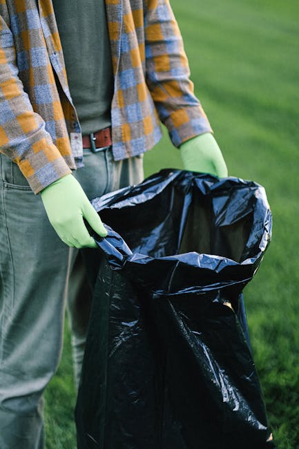 A person standing outdoors on a grassy area, dressed in a plaid shirt, grey trousers, and wearing light green gloves, is holding open a large black plastic rubbish bag with both hands. The bag appears